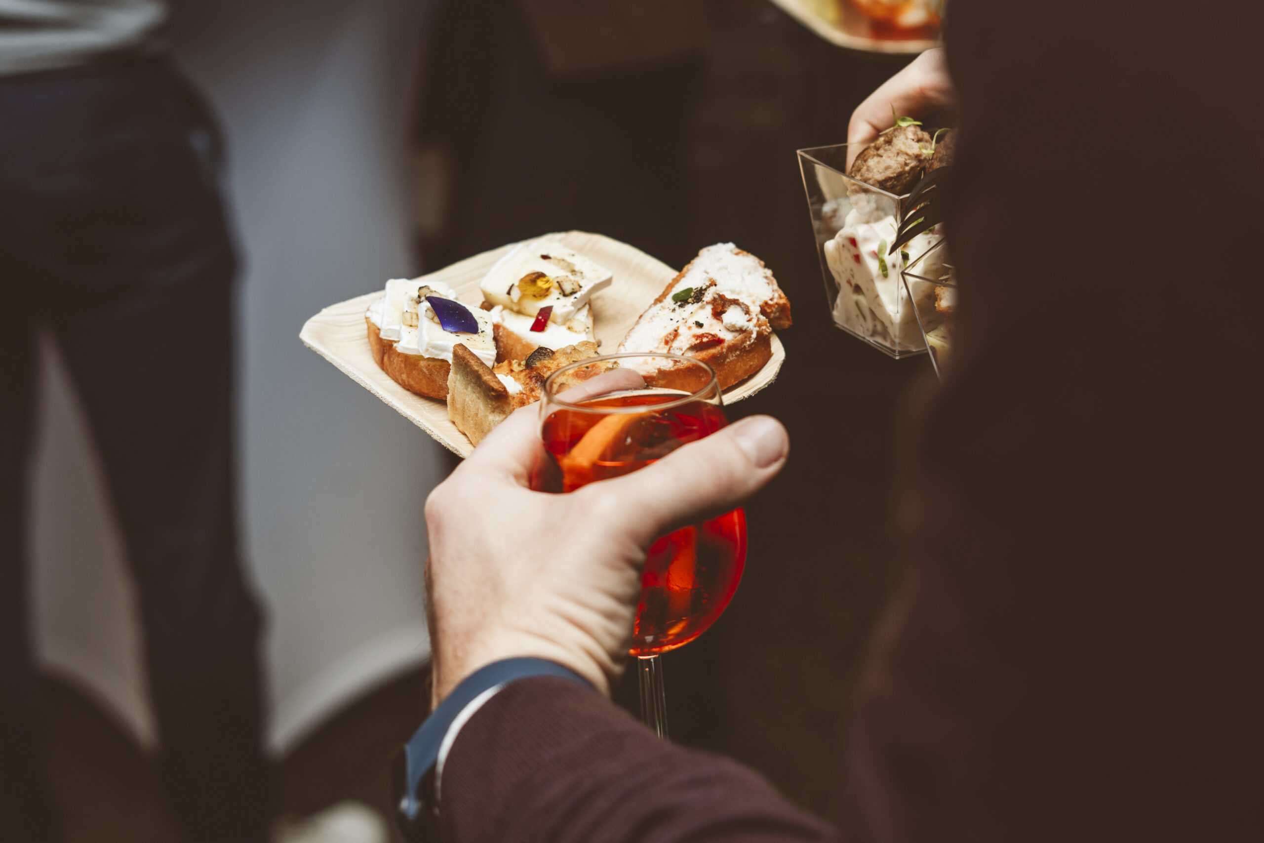Eine Person hält ein Glas Aperol Spritz und ein Teller mit Essen in den Händen | Fingerfood Catering in Hamburg
